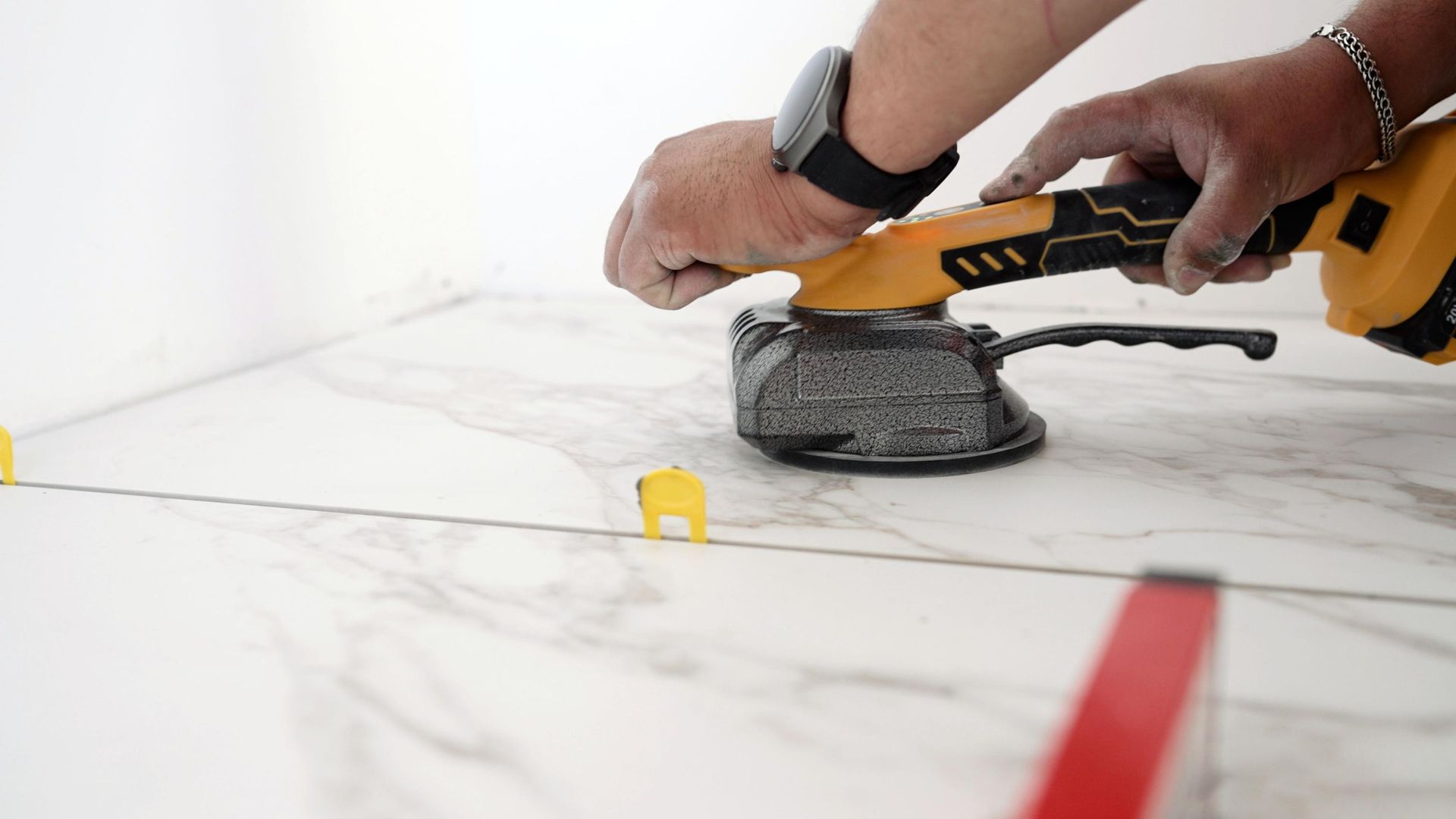 A person using a cordless vibrating suction cup tool to install large white marble-patterned floor tiles.
