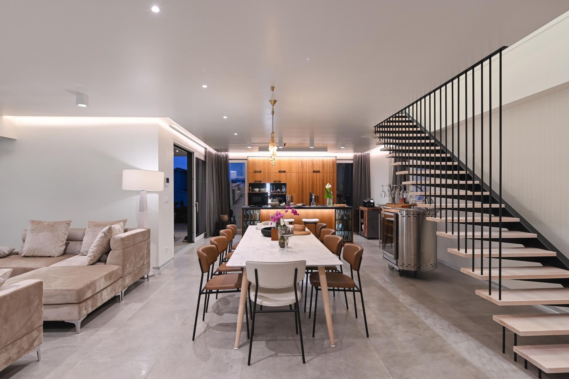 A modern, open-concept living and dining room with neutral furniture, a wooden kitchen, and an industrial staircase.