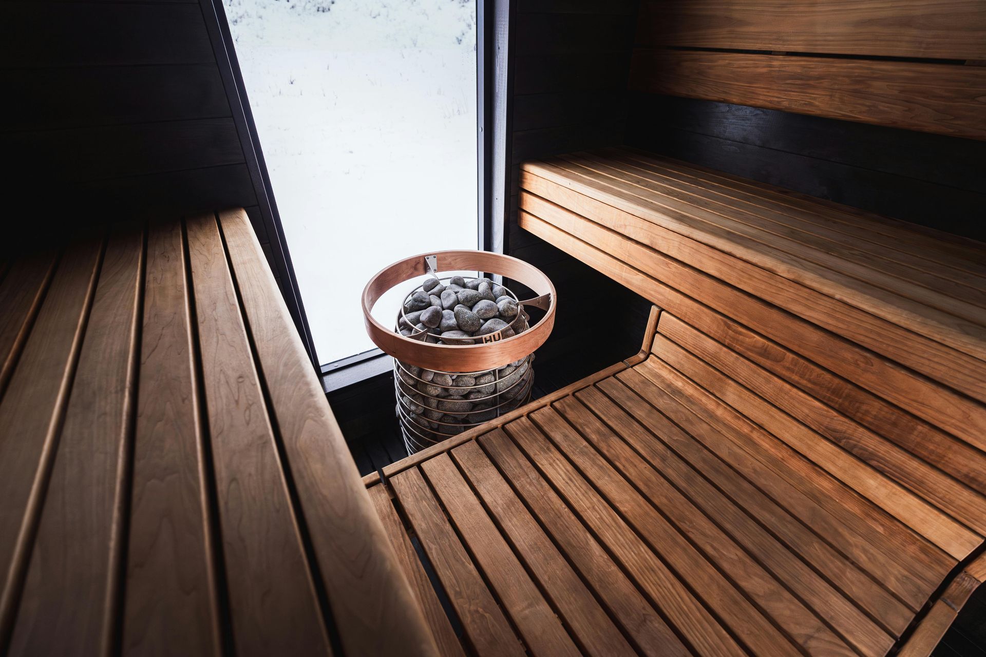 Wooden sauna interior with benches, a bucket of stones, and a window.