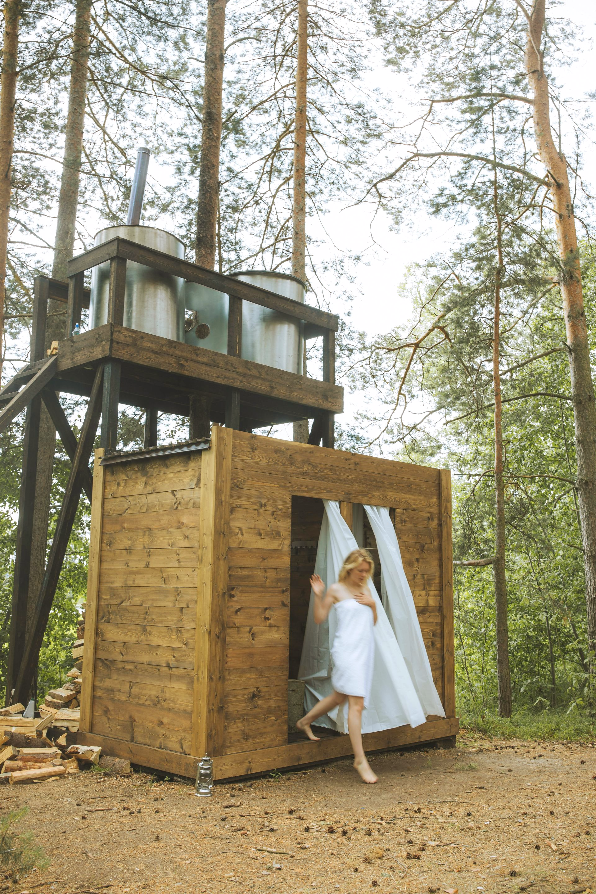 Woman in a white towel exits a wooden outdoor shower in a forest. A water tank is perched above.