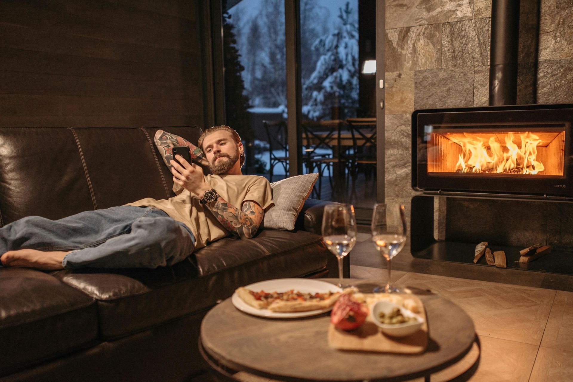 Man relaxing on a leather couch, looking at phone. Fireplace, table with food/wine, snowy outside view.
