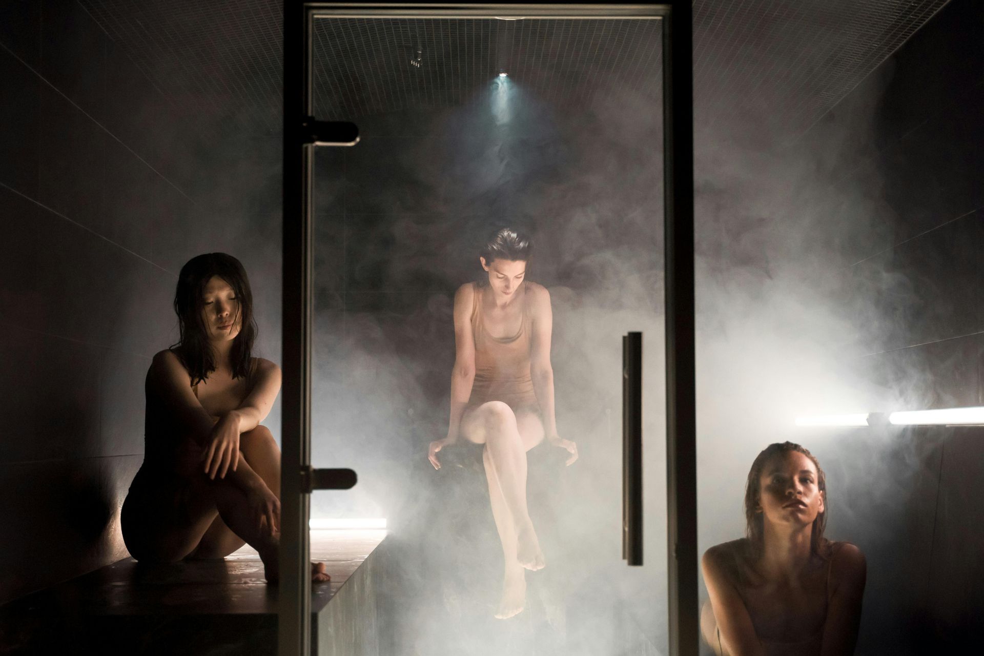 Three people in a steamy sauna: One sits, one is behind the glass, and another stands. Smoke fills the room.