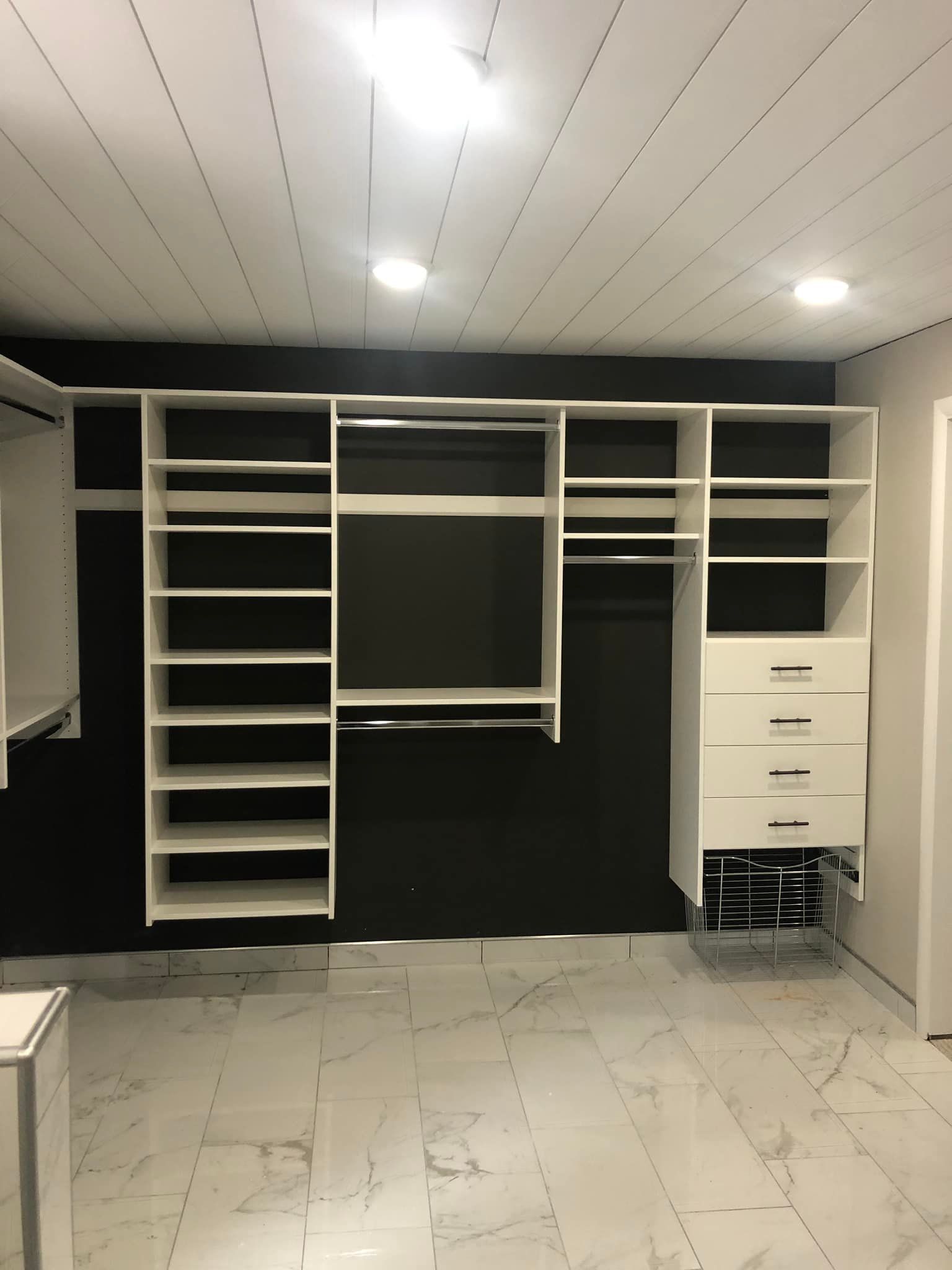 White custom closet with shelves, drawers, and hanging rods against a black wall. Marble floors.