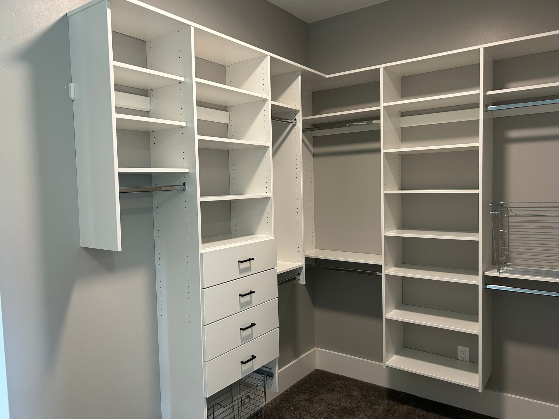 White closet organizers in a corner. Shelves, drawers, and hanging rods. Gray walls, dark carpet.