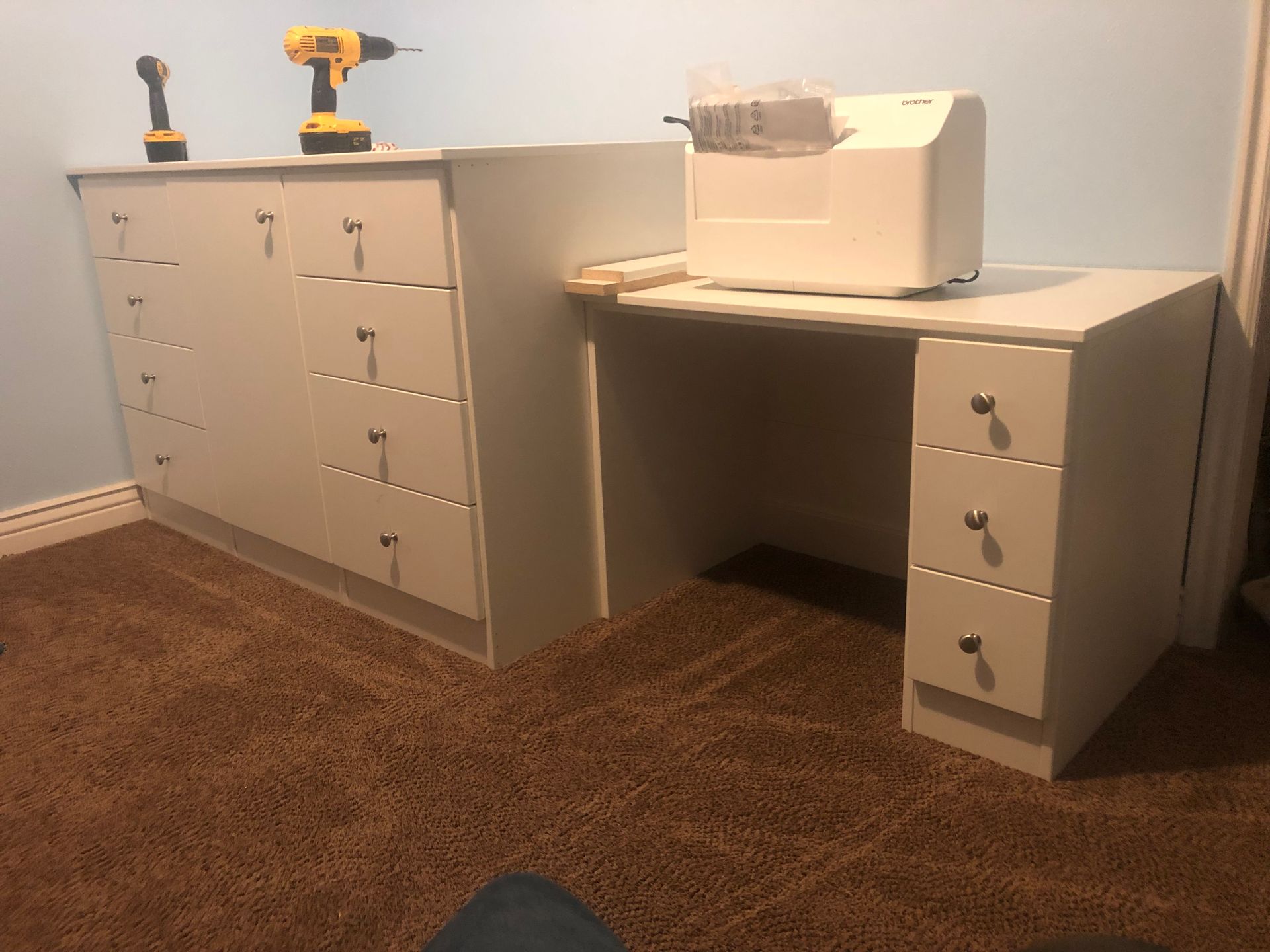 White desk with drawers, cabinet, and a sewing machine on a brown carpet.  A yellow drill and screwdriver are on top.