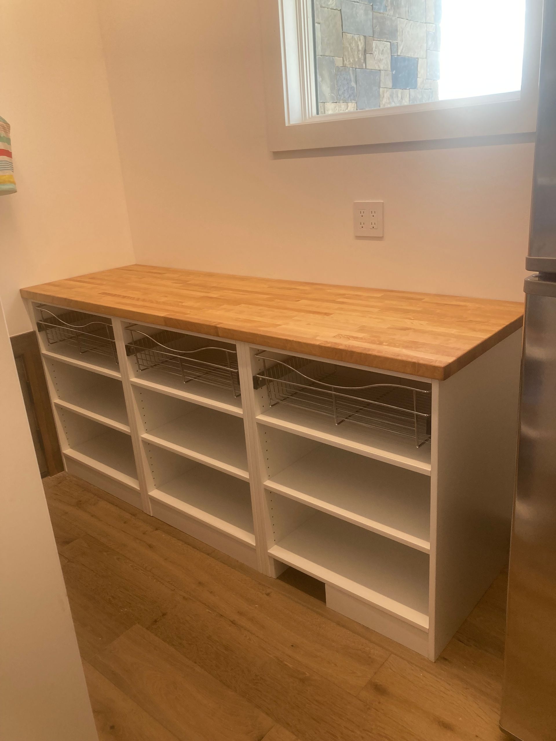 White shelving unit with wooden countertop, baskets, and shelves under a window.