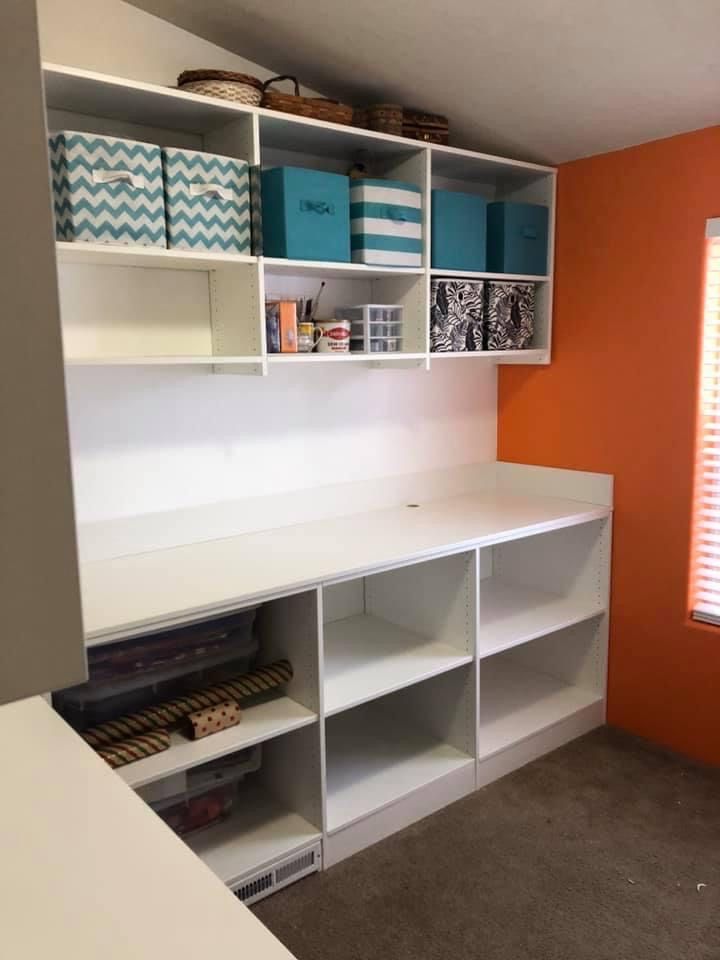 White shelving unit in room with orange wall; includes storage boxes, workspace, and rolls of paper.