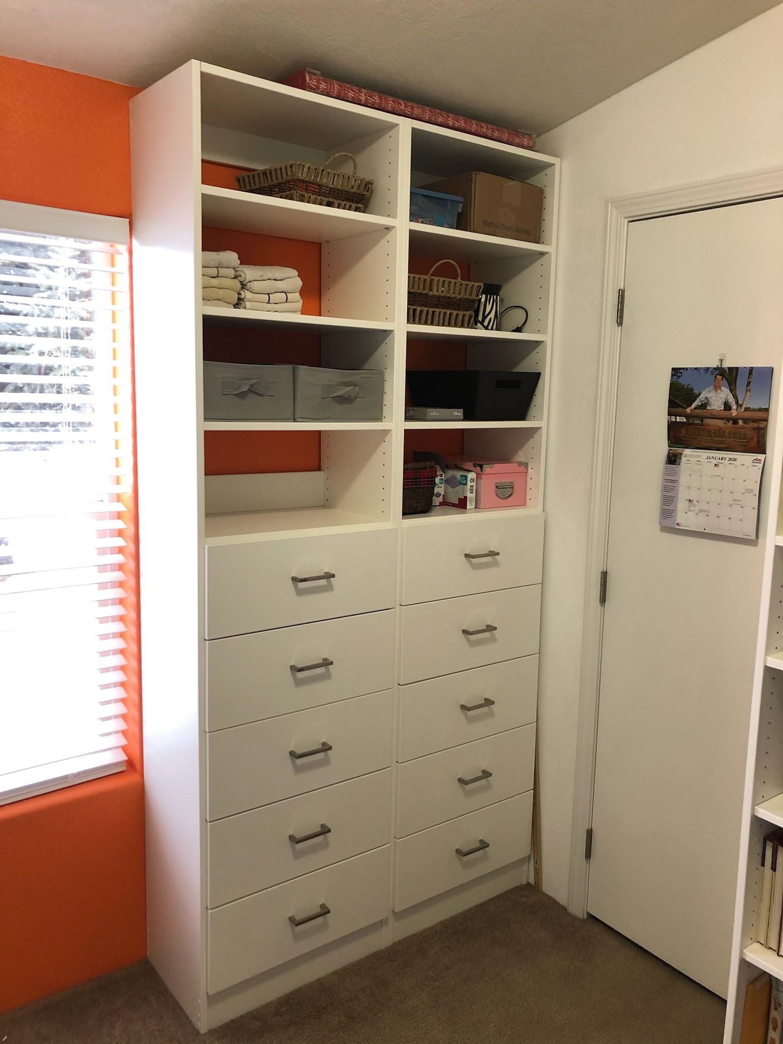 White storage unit with drawers and shelves next to an orange wall and a white door.