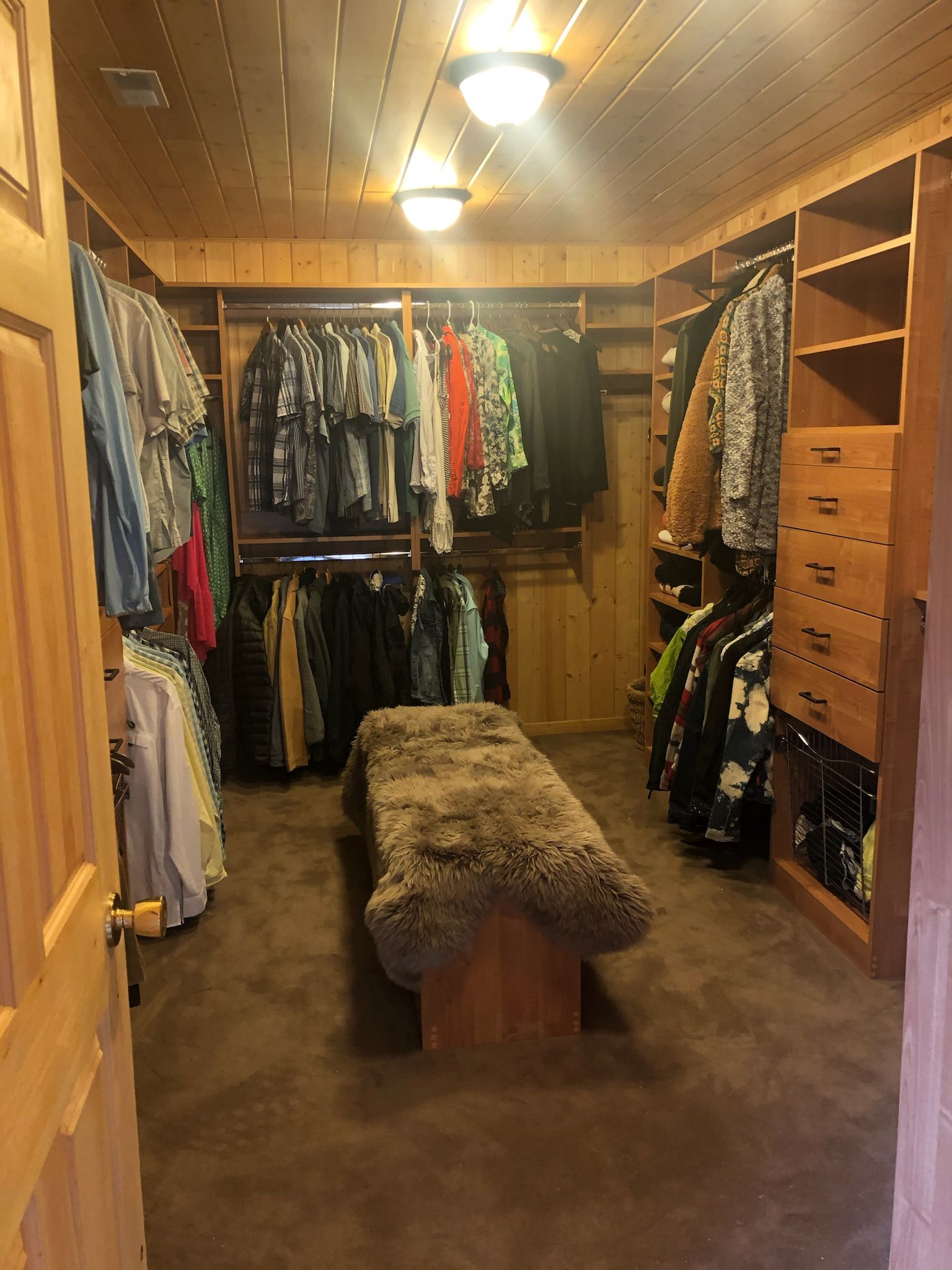 Walk-in closet with wooden shelves, a bench, and clothes hanging. Brown walls and flooring.