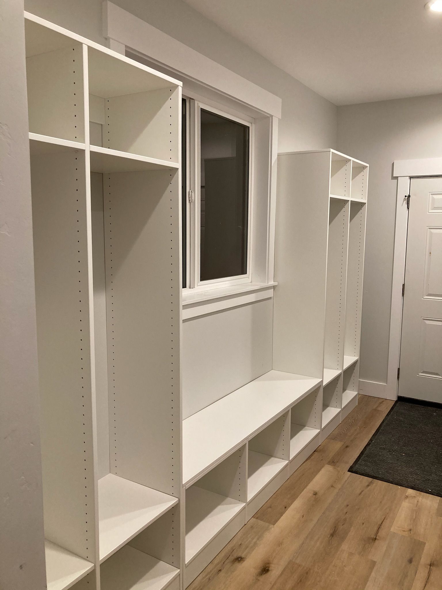 White built-in storage unit with shelves, bench, and window, beside door on wood floor.