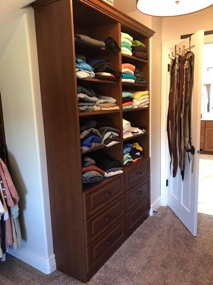 Wooden wardrobe with drawers and shelves holding clothes; belts hang on nearby door.