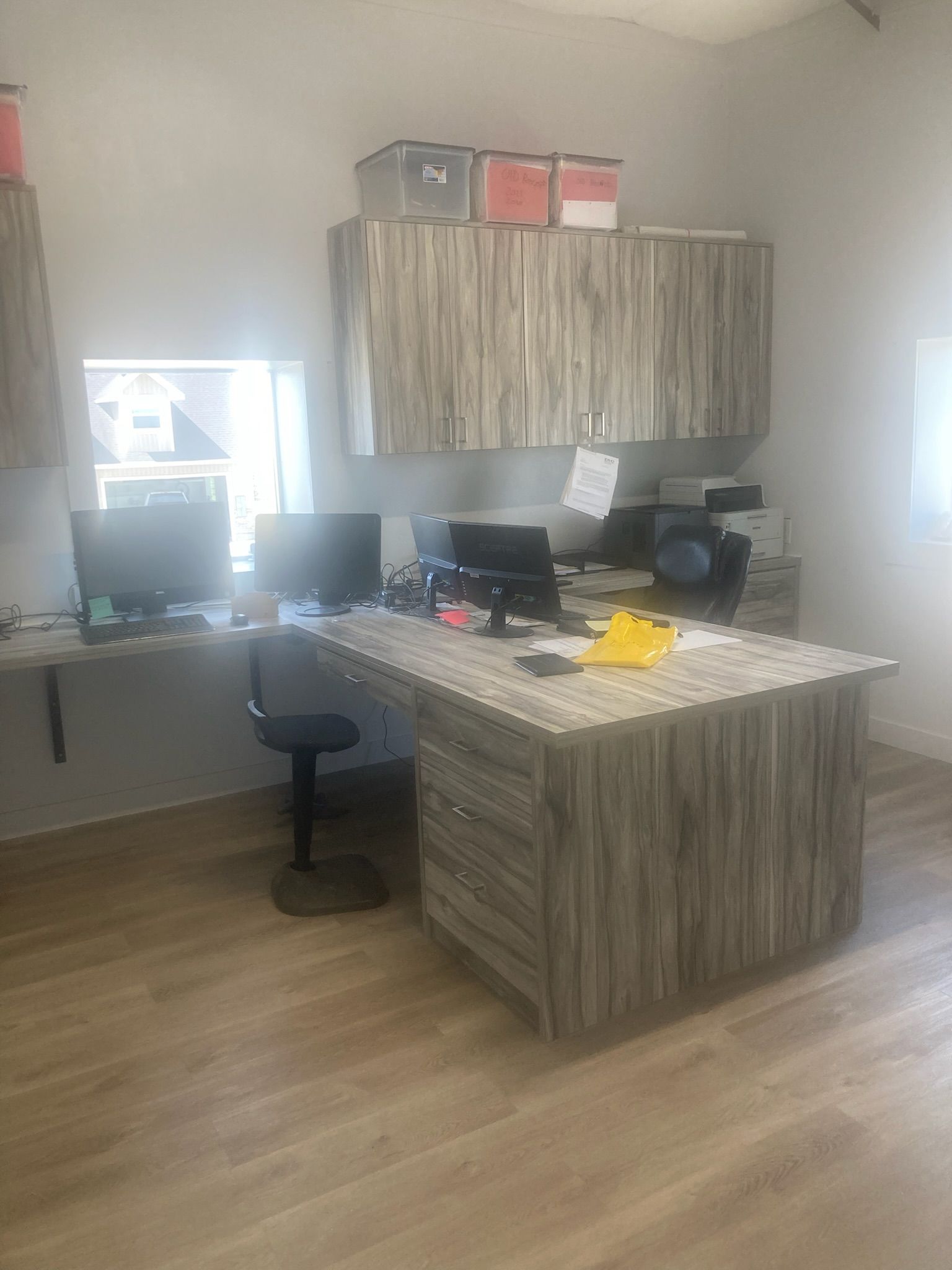 Office with wooden desk, cabinets, computers, and a chair. Light brown flooring and grey walls.