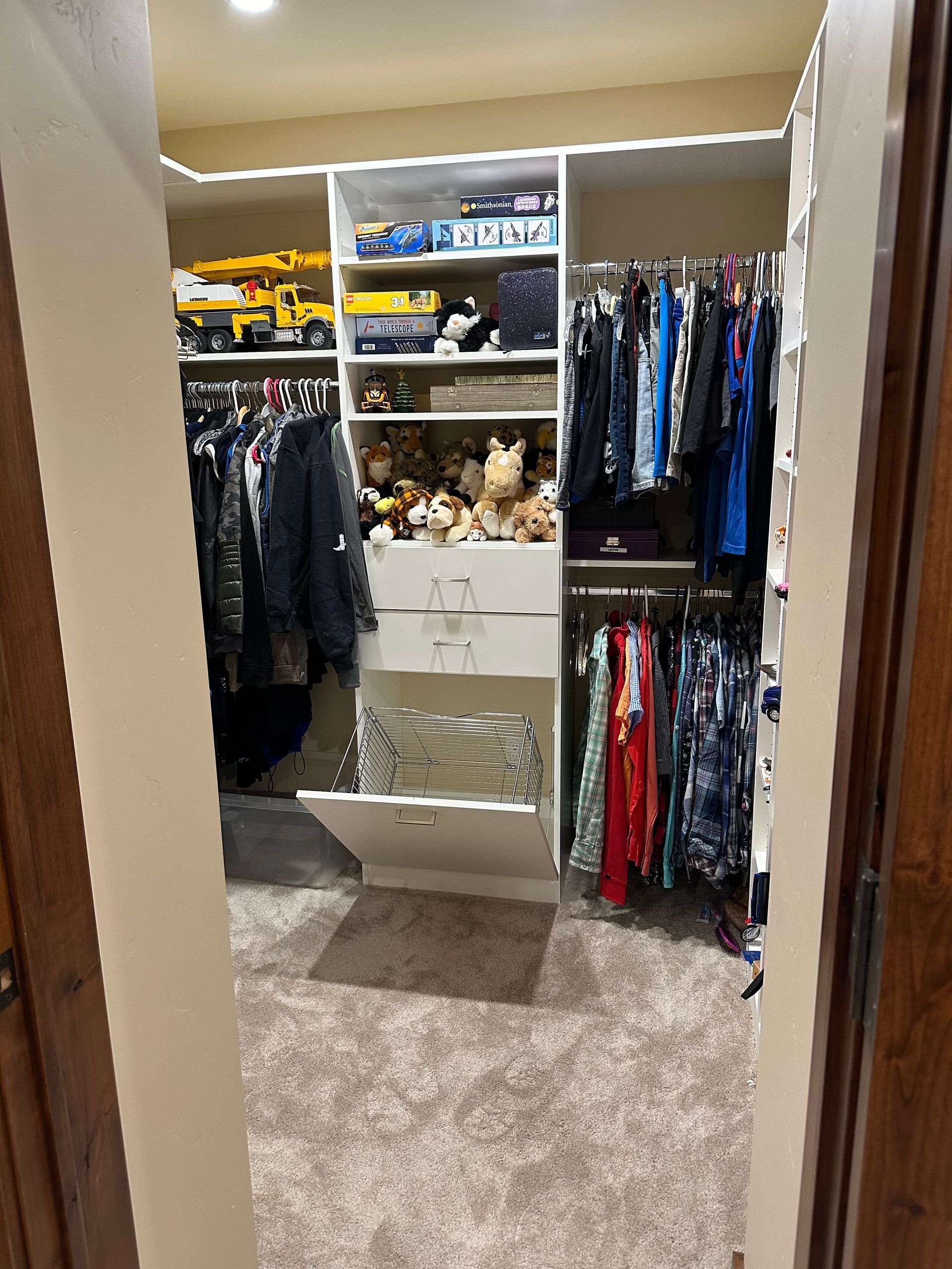 A walk-in closet with hanging clothes, shelving, and a dresser. Filled with items, it has tan carpet.