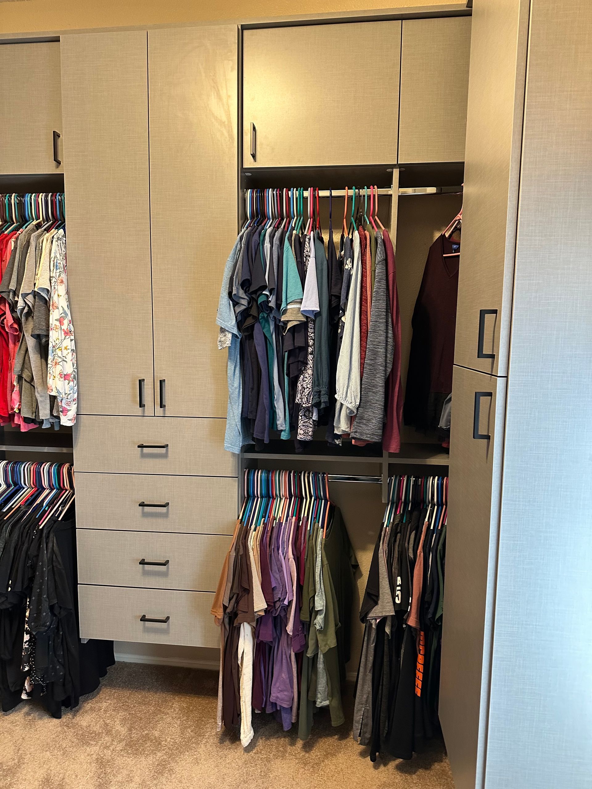 Walk-in closet with organized clothing. Tan cabinets, hanging clothes of various colors, drawers, and carpeted floor.