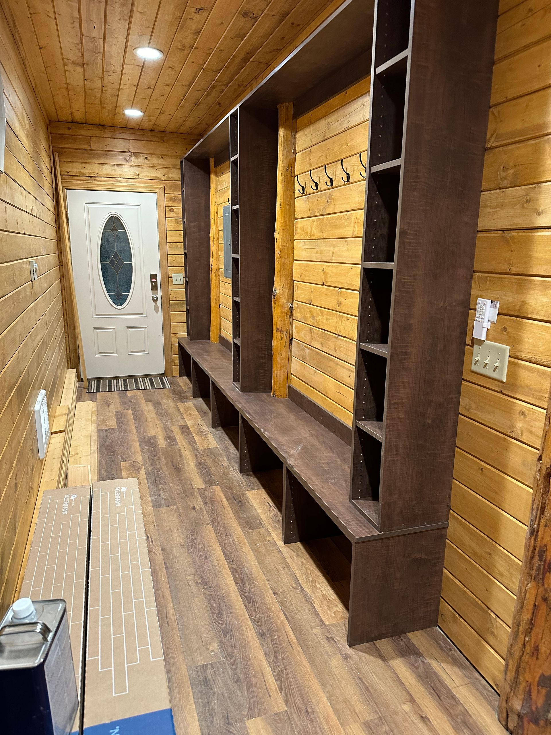 Hallway with wooden walls and floor. Brown shelving unit with bench and storage. White door at end.