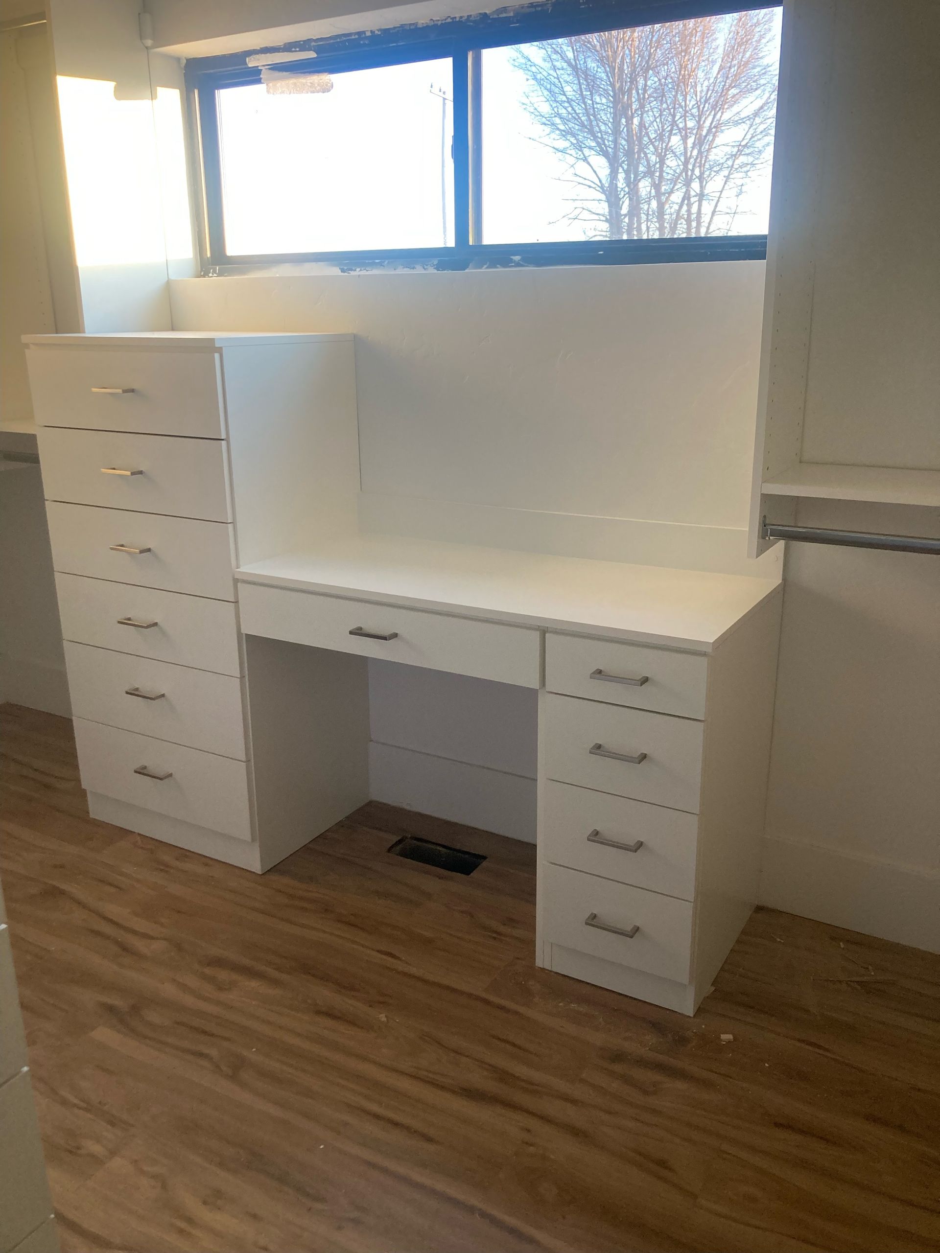 White vanity desk with drawers and a tall cabinet, against a window.