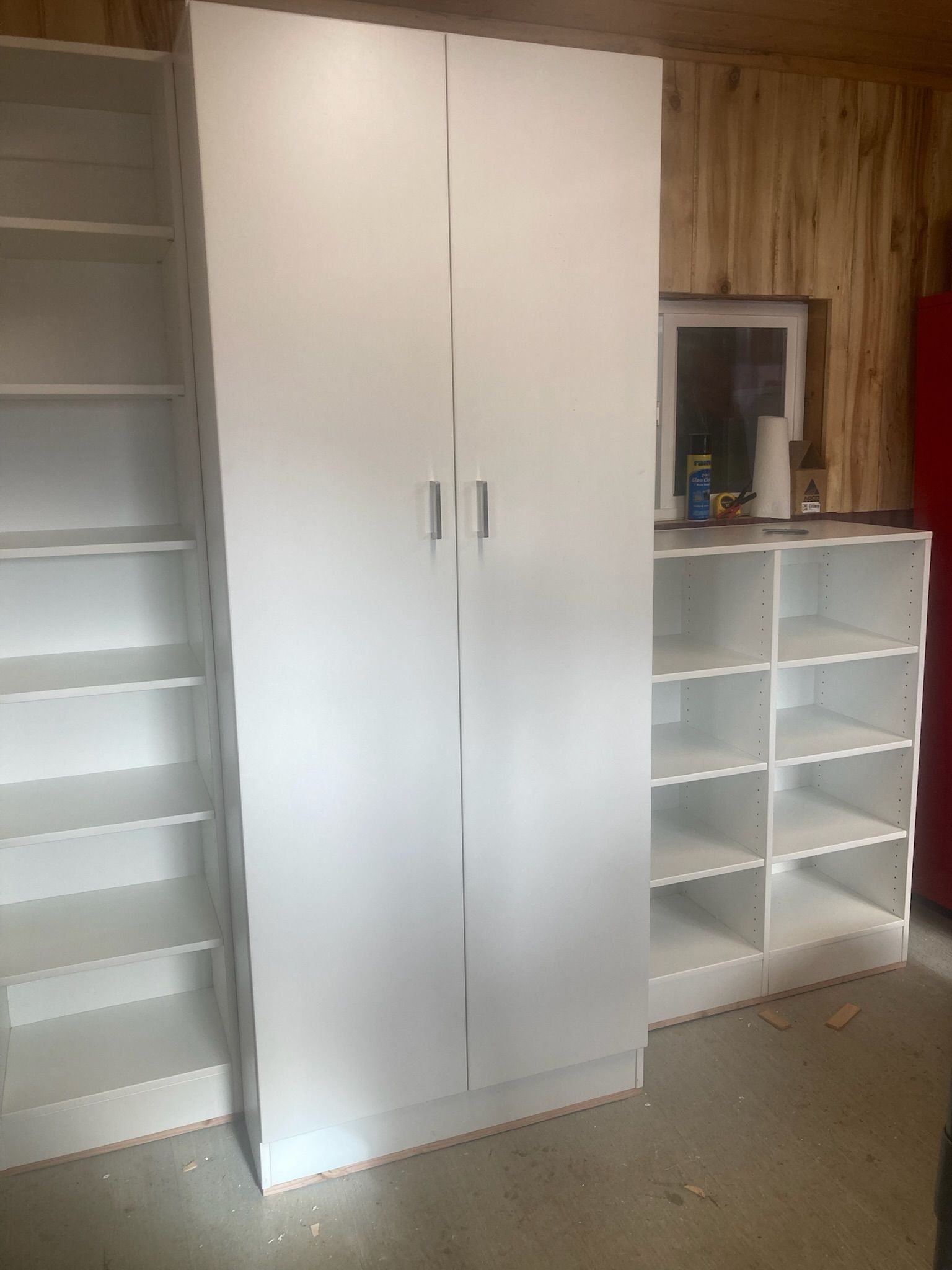 Three white storage units: tall cabinet, bookshelf, and cubby shelves against a wooden wall.
