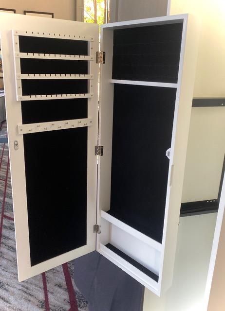 Open white jewelry armoire with black velvet lining, holding necklace hooks and shelf.