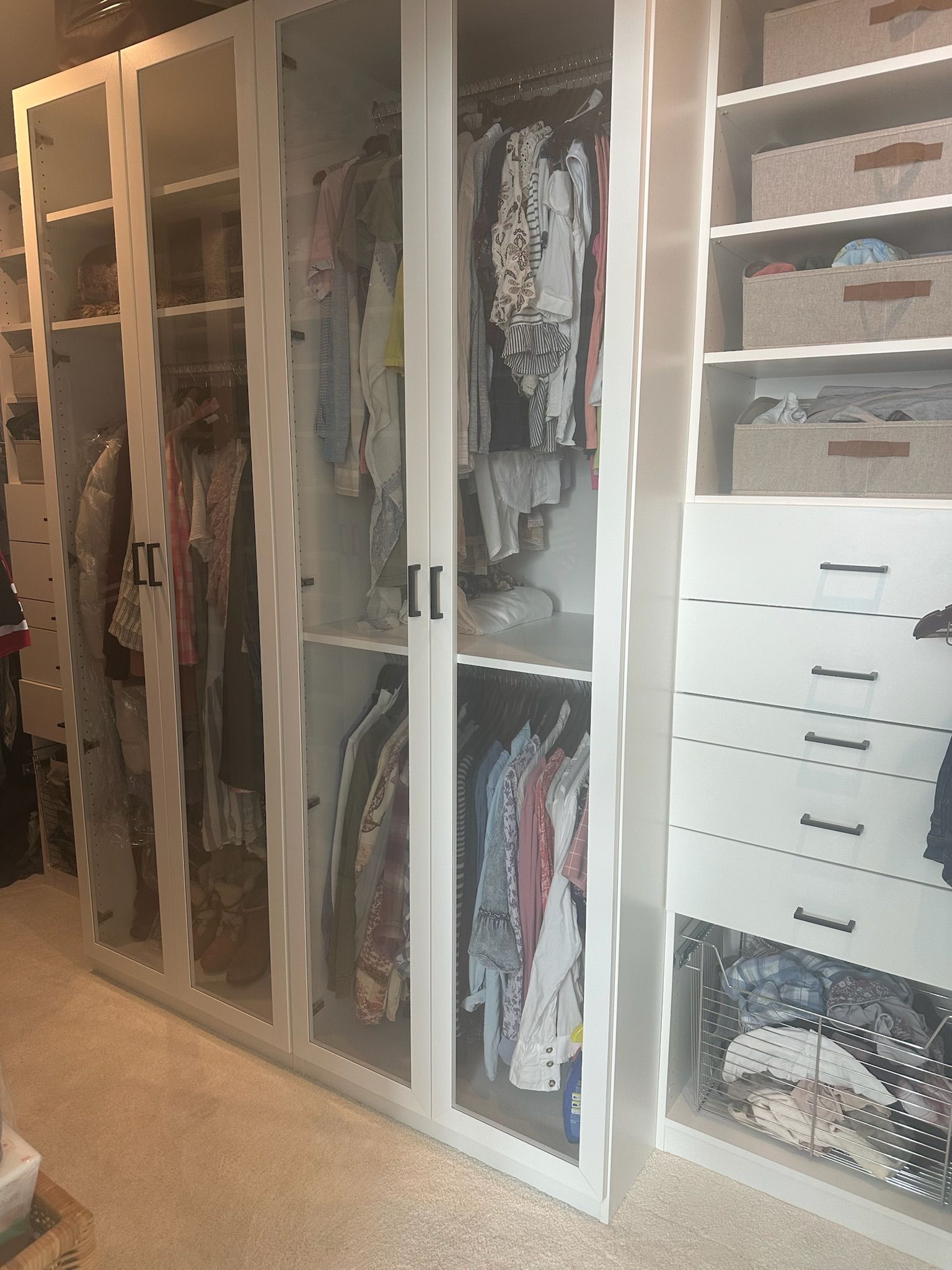White closet with glass doors, displaying clothes on hangers and drawers.