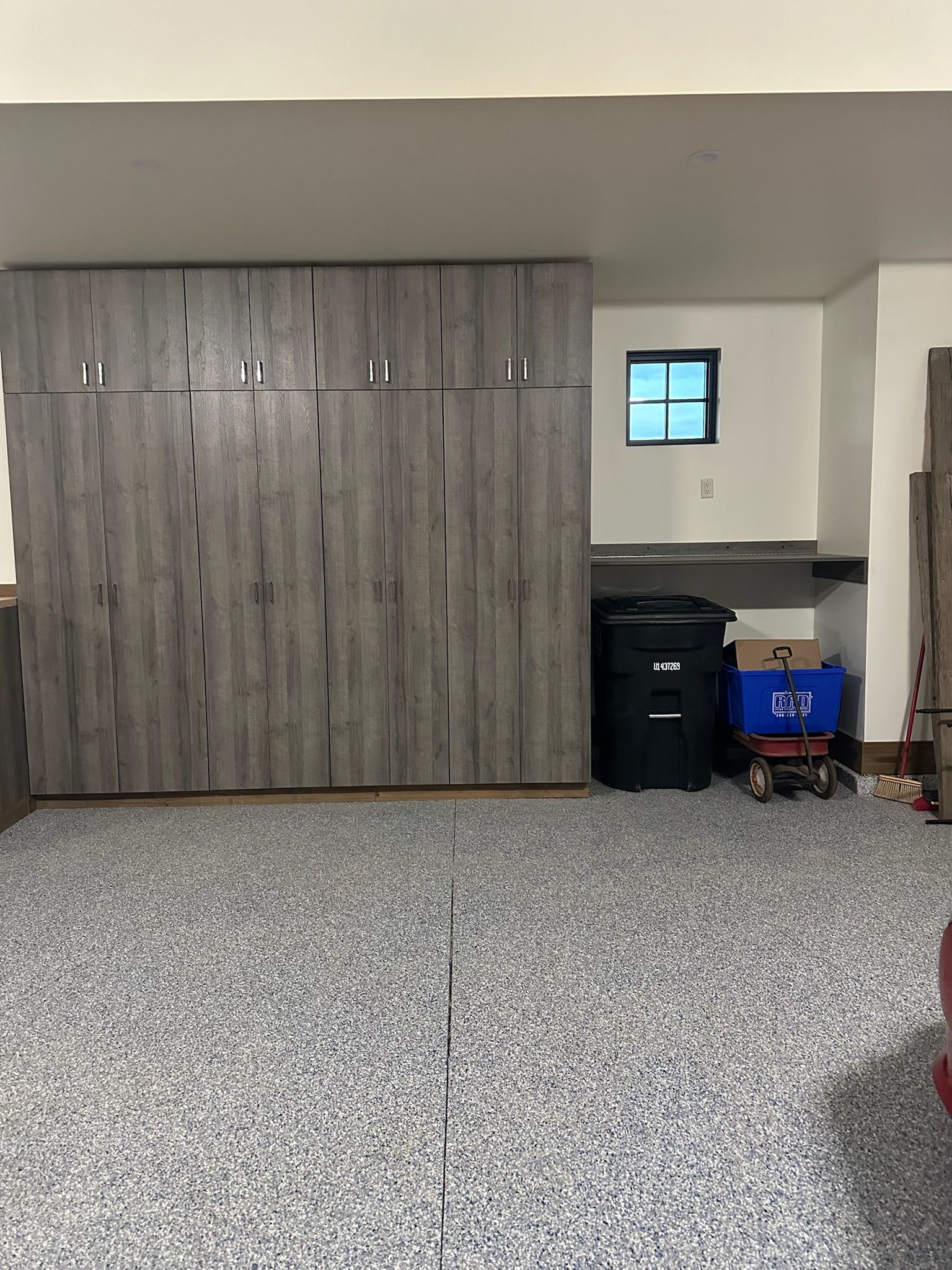 Garage with gray cabinets and speckled flooring. Black trash can, window, and red wagon visible.