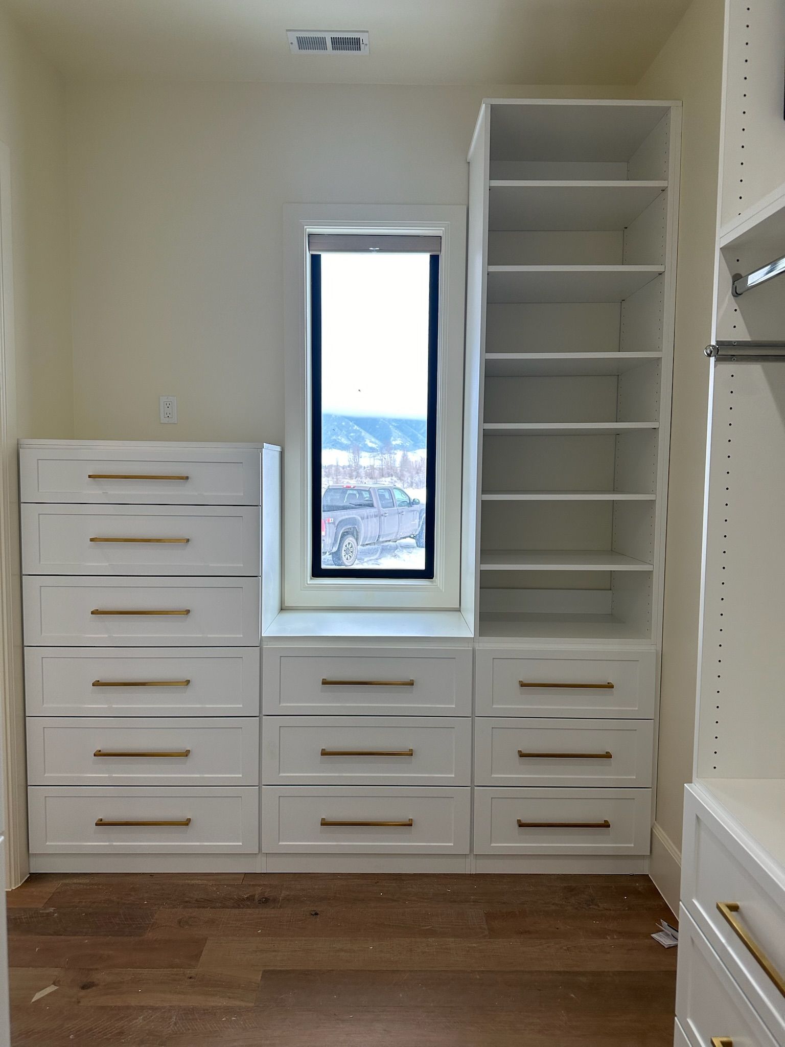 White closet with drawers, shelves, and a window. Gold hardware. Brown floor, beige walls.