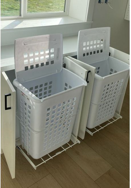 Two white laundry hampers on pull-out rails inside a cabinet, under a window.