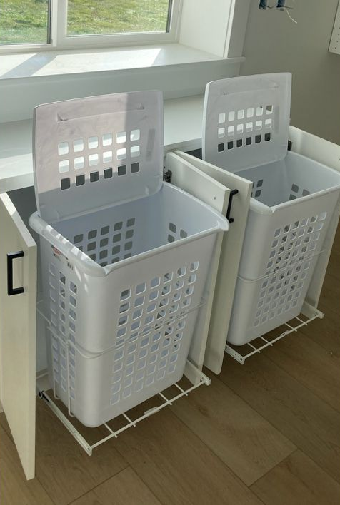 Two white laundry hampers on pull-out wire racks inside a cabinet, next to a window.