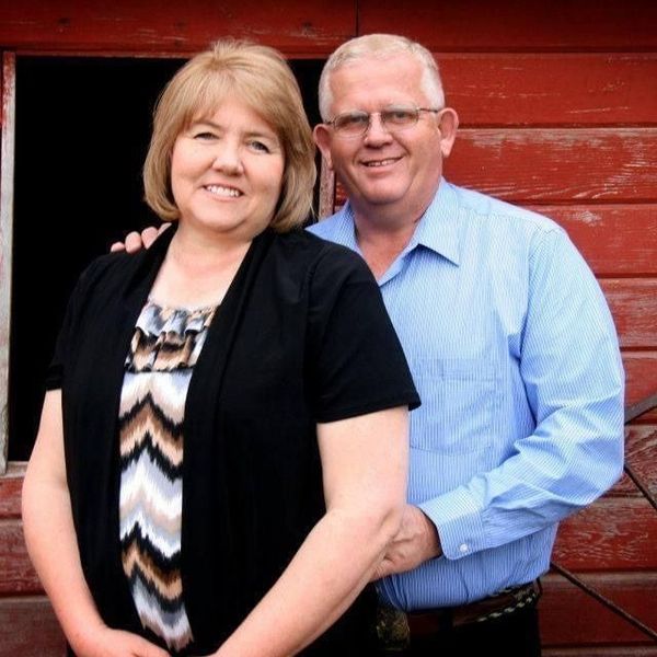 Smiling couple, woman in black cardigan, man in blue shirt, leaning against a red barn.