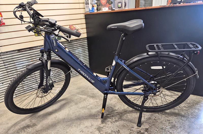 A dark blue step-through electric bicycle with a rear rack and suspension fork, parked indoors on a concrete floor.