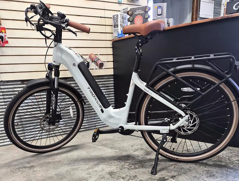 A white Troxus e-bike with tan grips and seat, black tires, and a rear rack, standing on a concrete floor in a shop.