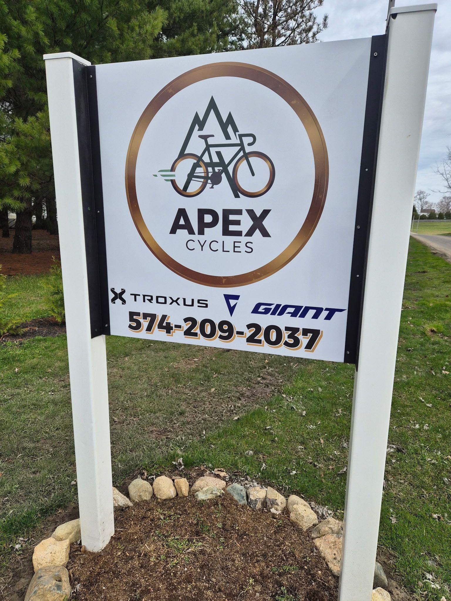 A sign for Apex Cycles featuring a bicycle and mountain graphic, brand logos, and the phone number 574-209-2037.