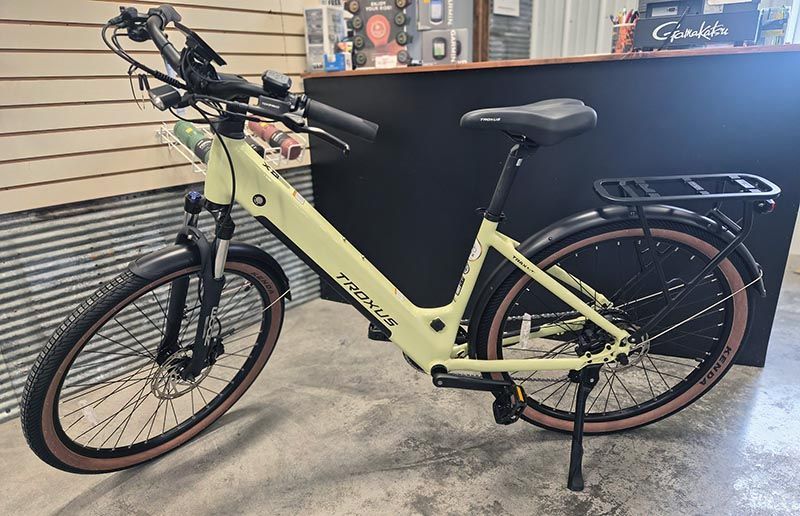 A pale yellow electric step-through bike with a black rear rack, fenders, and knobby tan-wall tires in a shop.