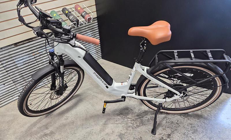 A white electric step-through bicycle with a brown seat and grips, parked on a concrete floor in front of a slatted wall.