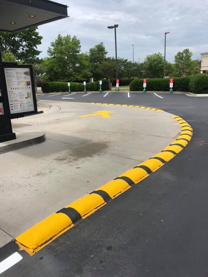 A yellow and black striped curb barrier curves along a drive-thru lane leading to a menu board and parking spaces.