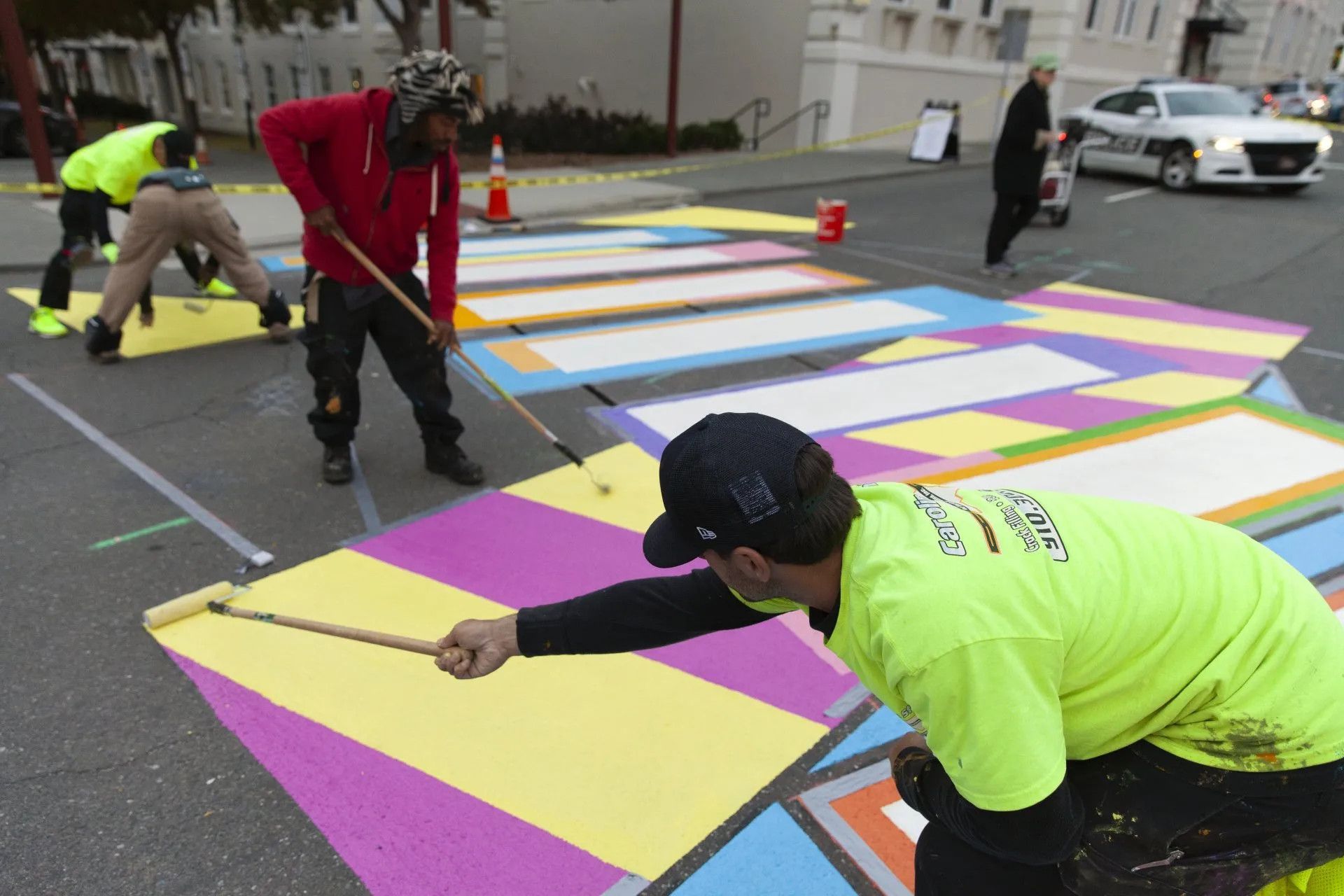 Workers in high-visibility clothing paint a colorful, geometric mural across an asphalt street crosswalk.
