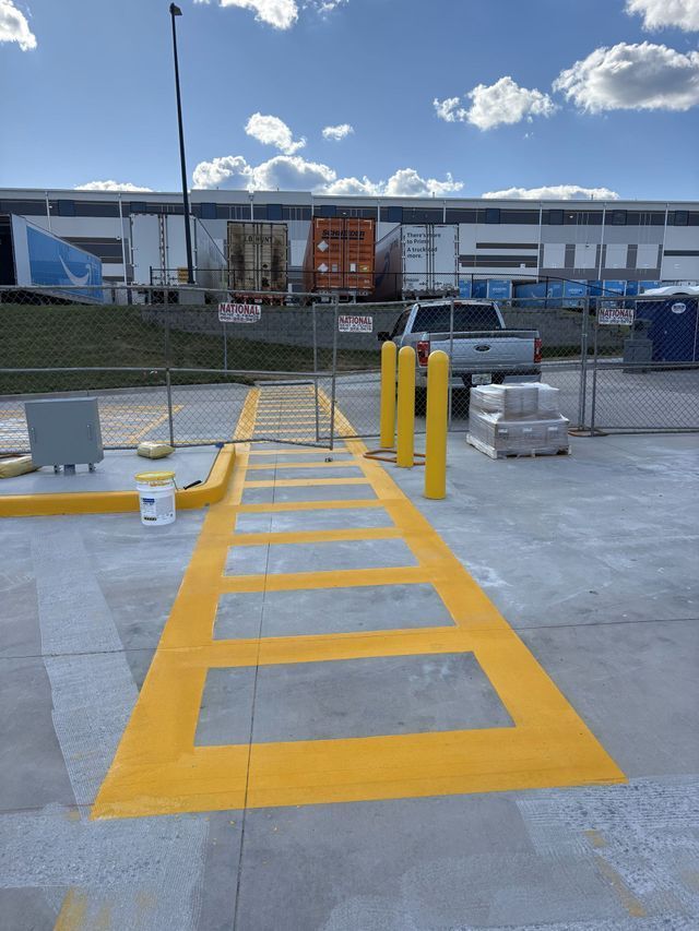 A yellow painted pedestrian walkway leading toward a fenced warehouse loading dock and a pickup truck.