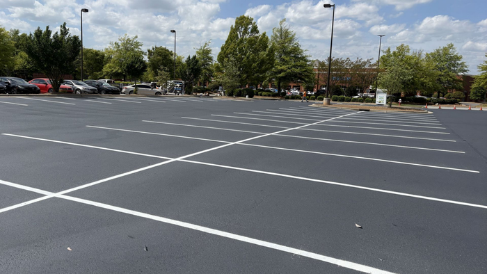 A newly paved, empty parking lot with clearly marked white parking stalls, surrounded by green trees under a cloudy sky.