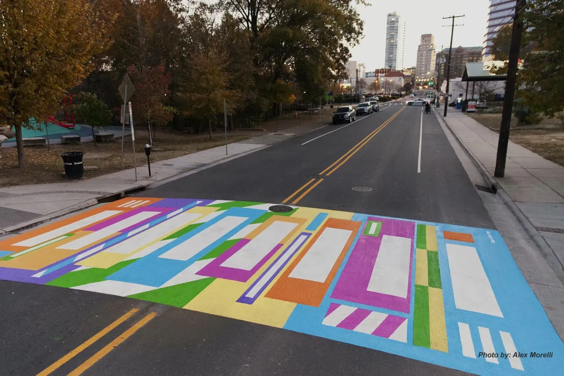 A colorful, abstract-patterned pedestrian crosswalk on an asphalt road in a city street setting.