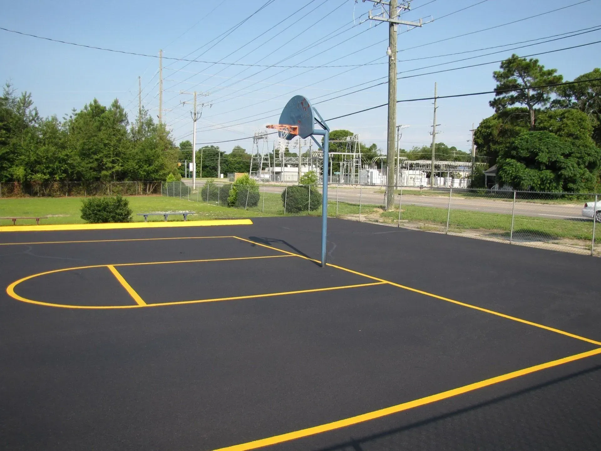 A basketball hoop with a blue backboard stands on a freshly paved outdoor court with yellow painted lines.