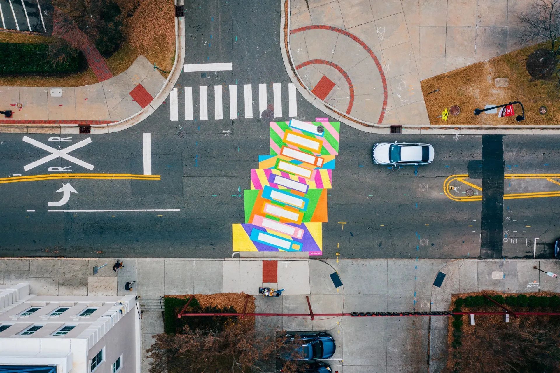 An aerial view of a vibrant, multi-colored crosswalk at a city intersection with a car driving across it.