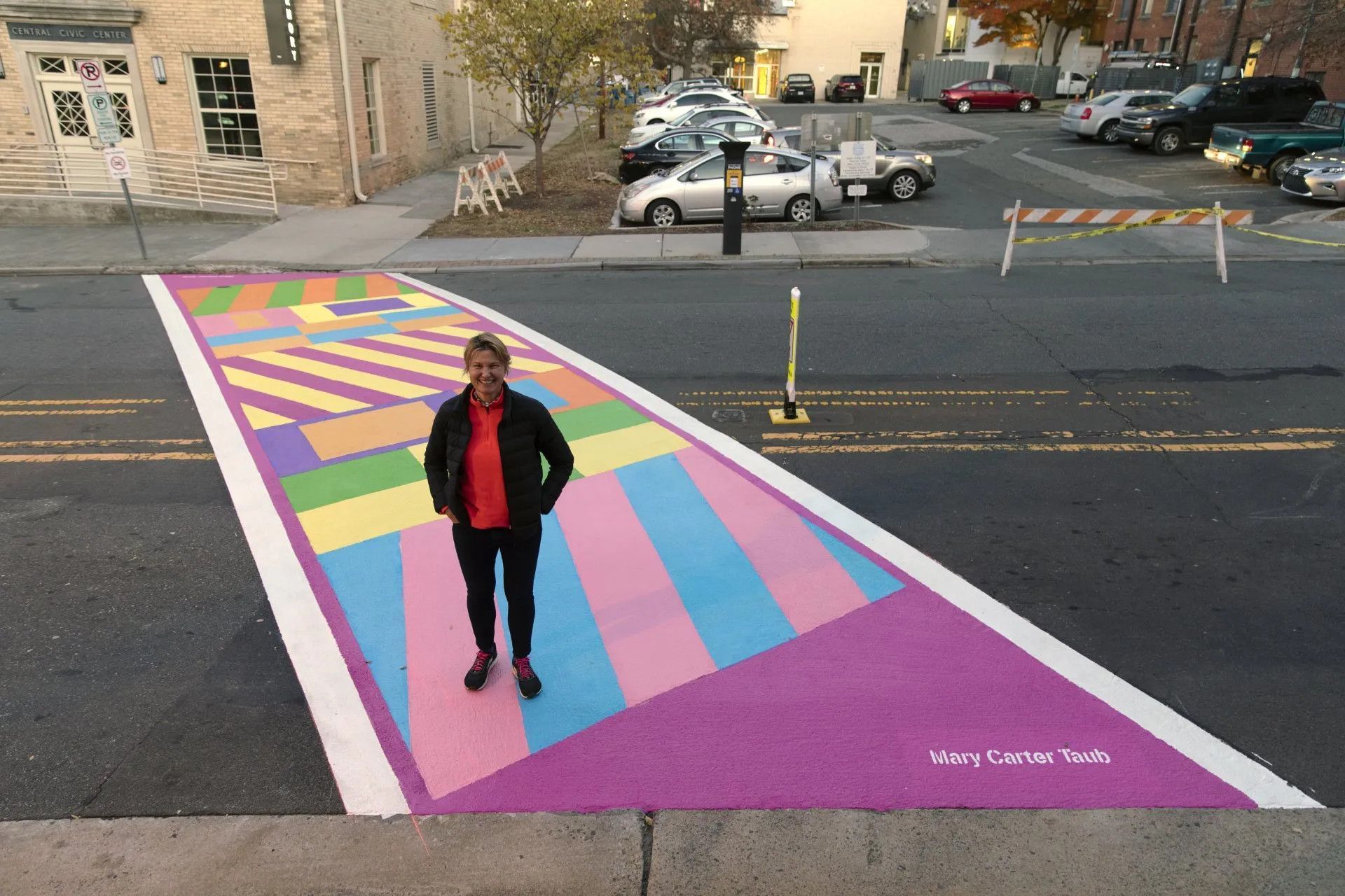 A person stands on a multi-colored geometric mural painted across a street crosswalk in an urban setting.