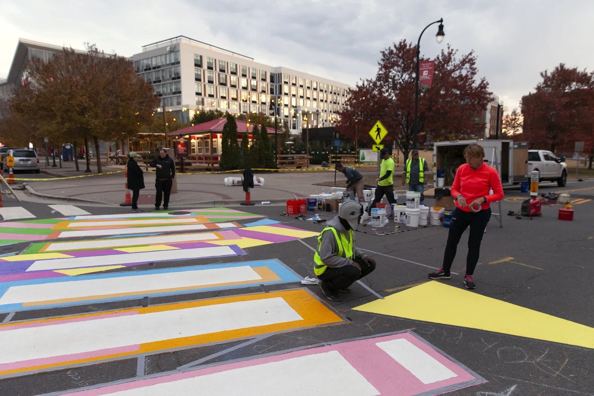 Volunteers wearing safety vests paint a colorful crosswalk on a paved street near a large building.