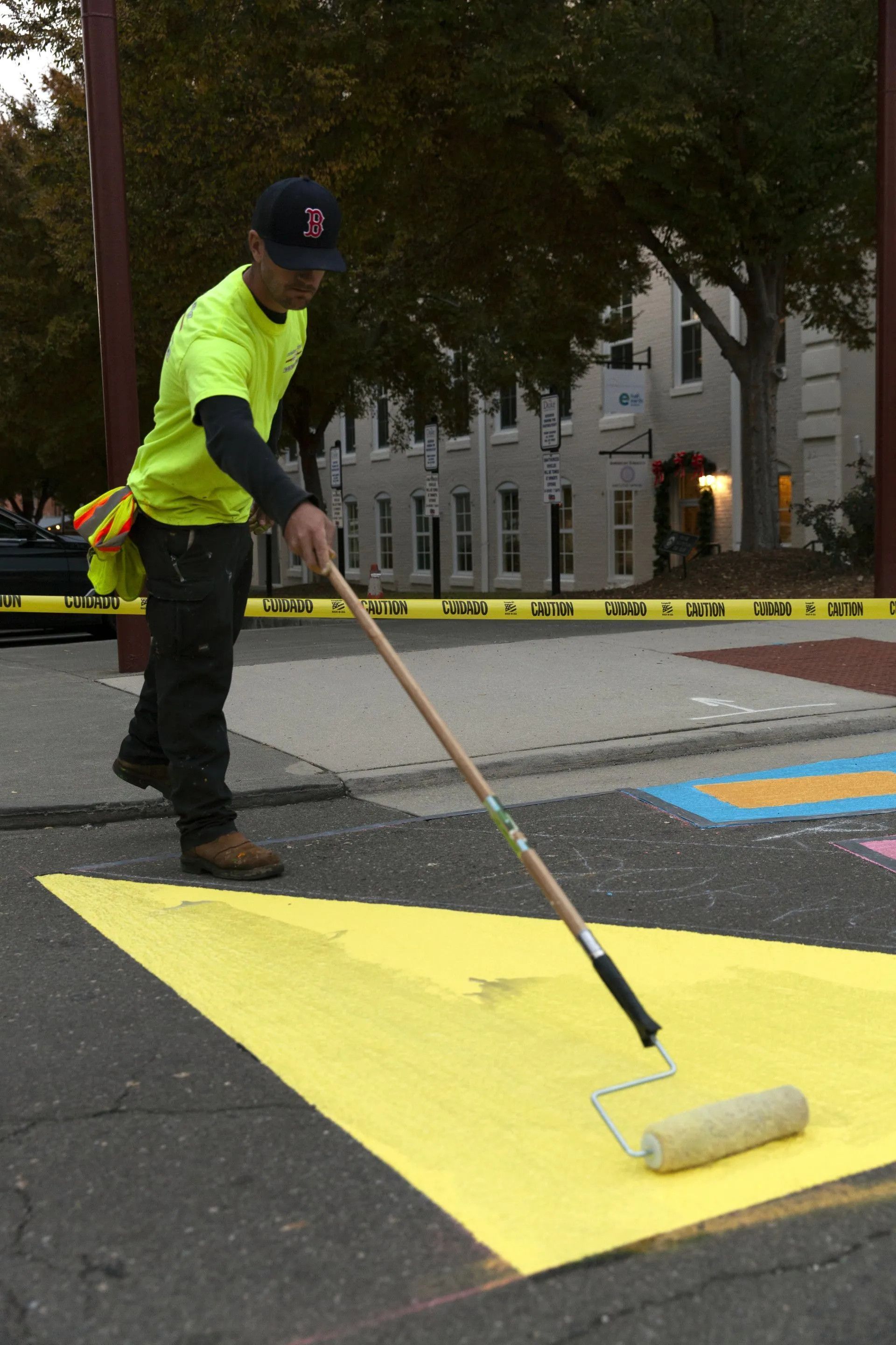 A worker in a bright yellow shirt uses a paint roller to apply yellow paint to a geometric shape on an asphalt street.