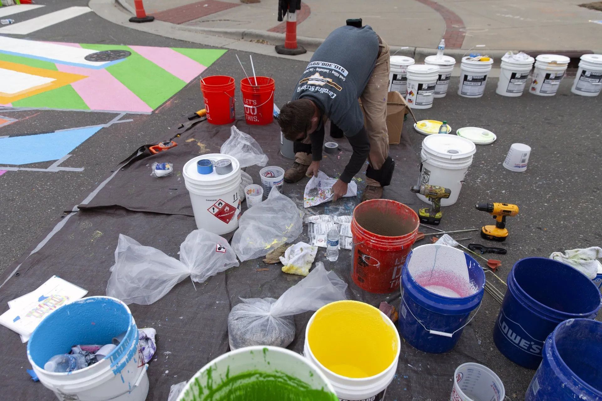 A person crouches on an asphalt surface, painting a colorful street mural, surrounded by numerous buckets of paint.