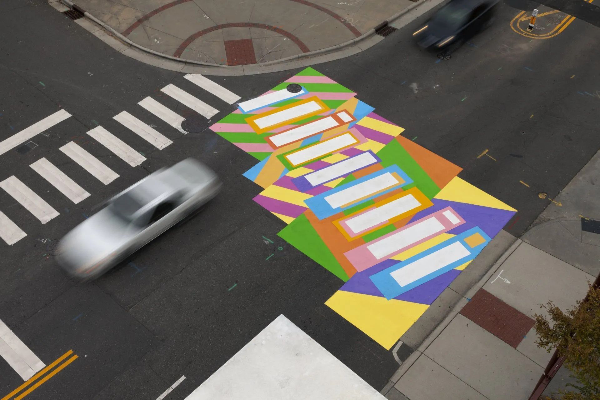 An aerial view of a colorful, geometric street mural surrounding a white pedestrian crosswalk at a city intersection.