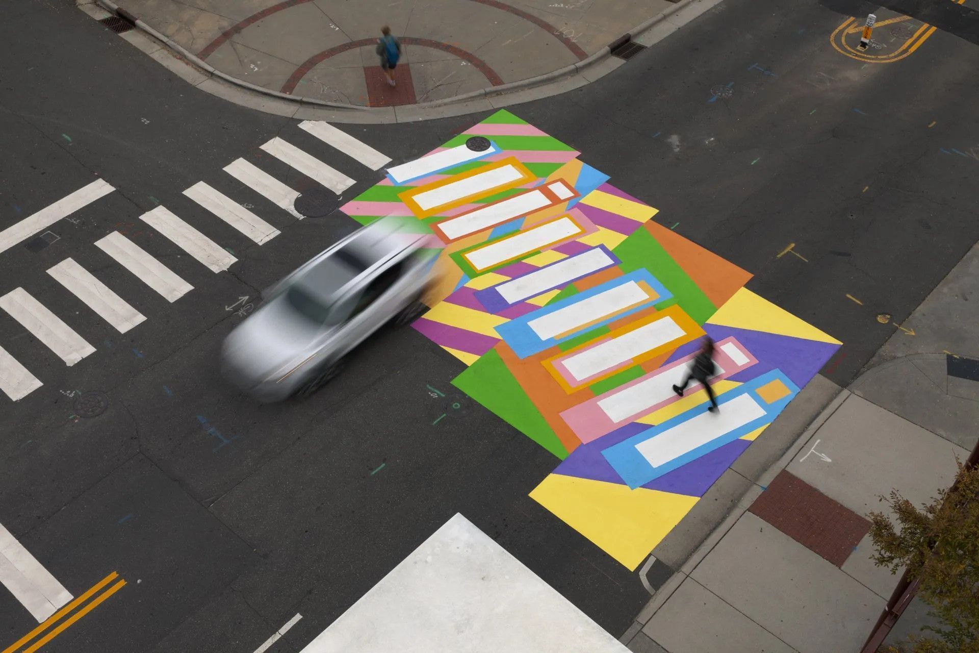 An aerial view of a pedestrian crossing painted with colorful geometric patterns, with a car and a person walking across.