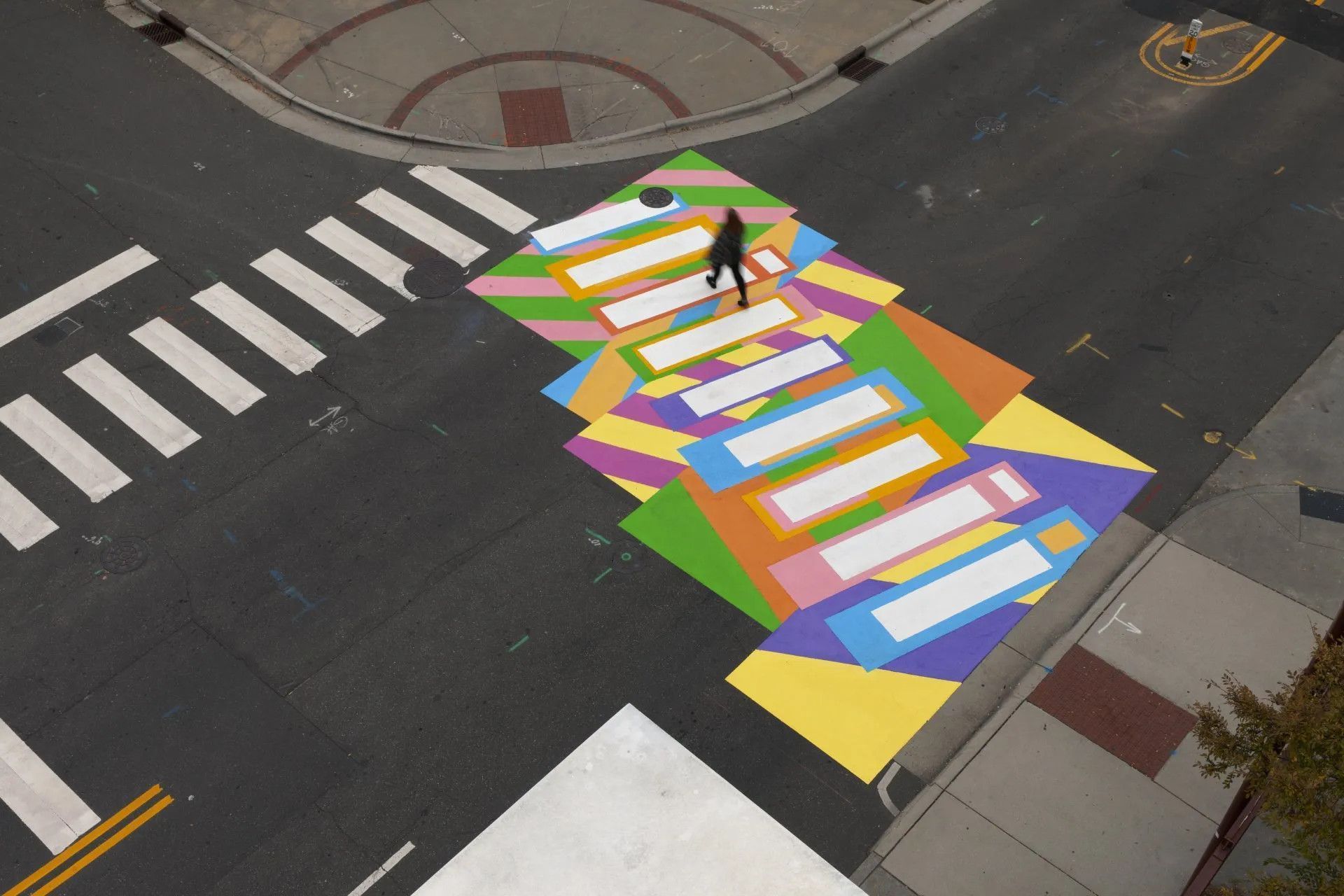 An aerial view of a pedestrian crossing painted with colorful geometric patterns, with a person walking across it.