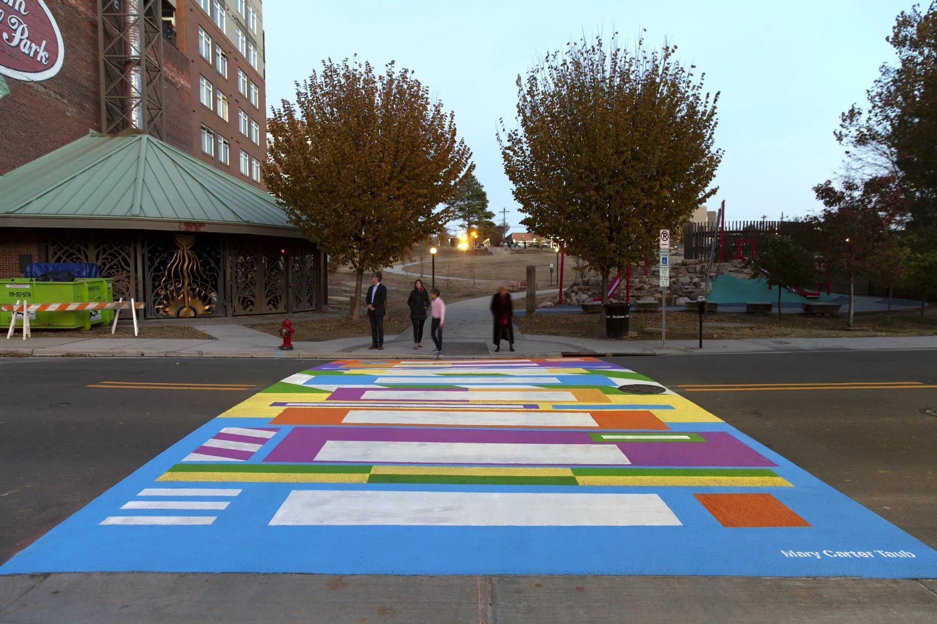 A colorful, geometric mural painted across a street as a pedestrian crosswalk, with three people standing on the sidewalk.