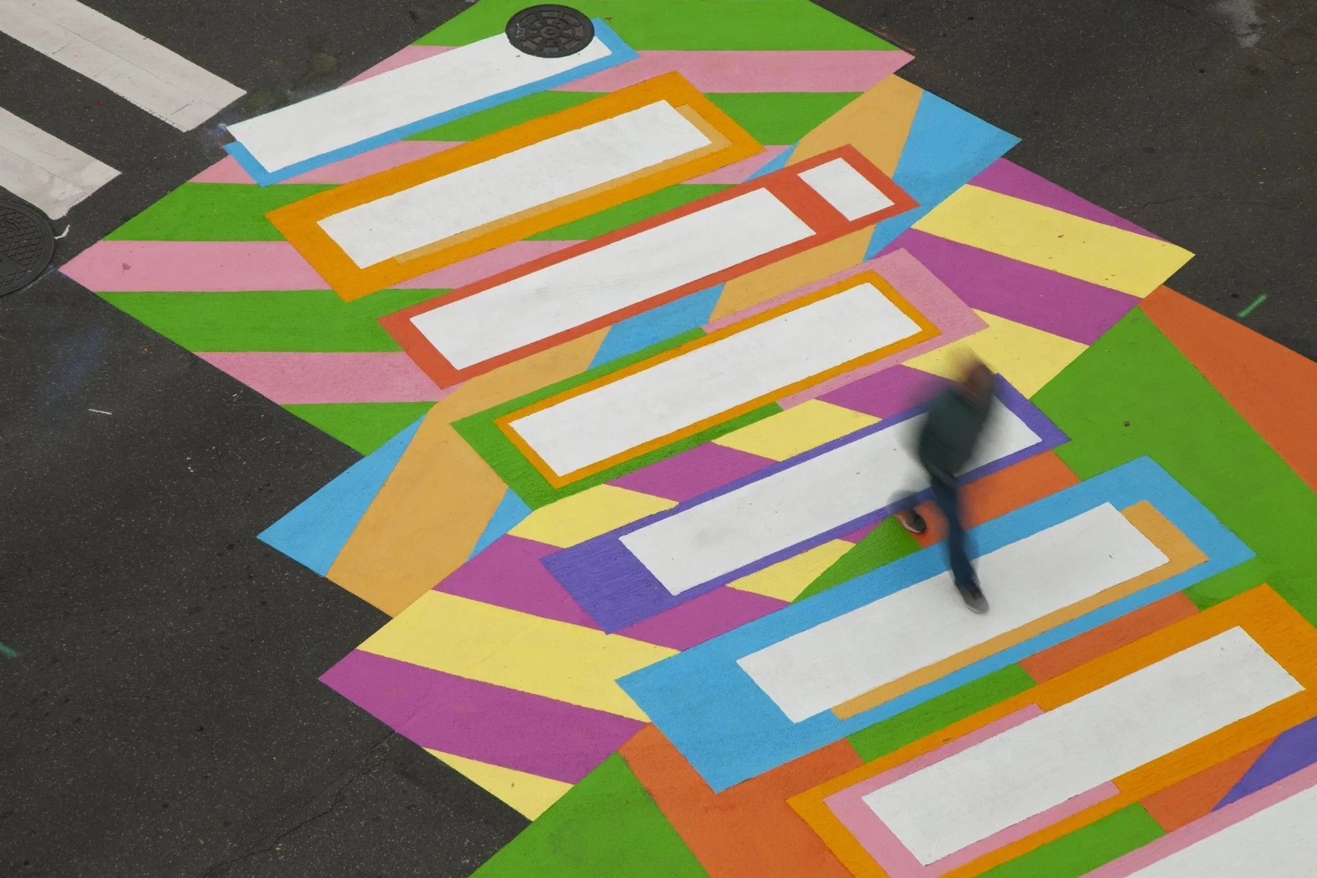 A person walks across a vibrant, multi-colored crosswalk painted with bright stripes and rectangular white bars.