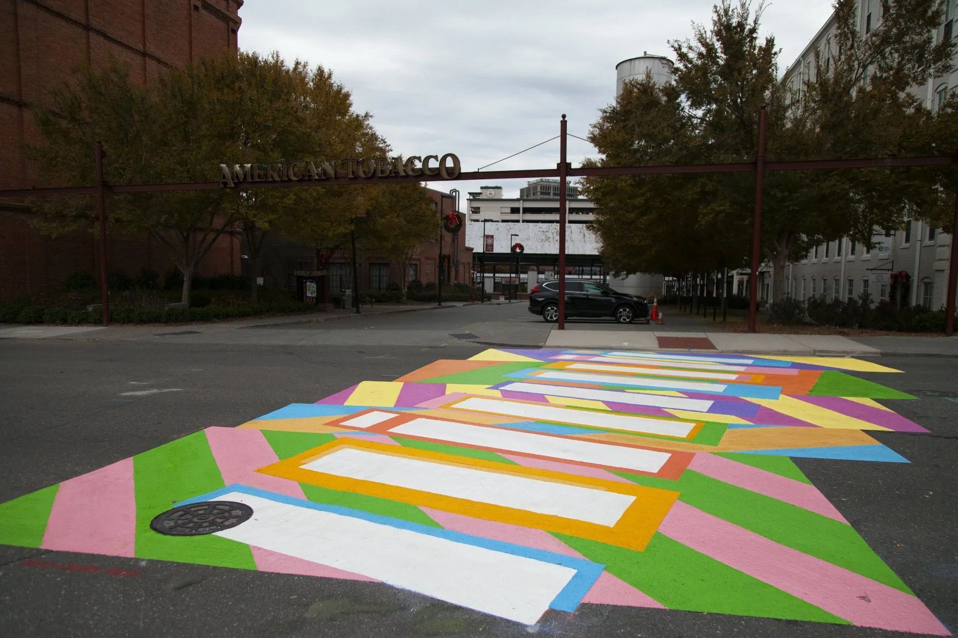 A colorful, geometric mural painted across an asphalt street with trees and industrial buildings in the background.